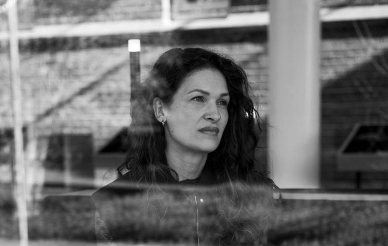 Vrouw zit op een bankje achter een raam, kijkt naar buiten in gedachten verzonken met een schrift op schoot