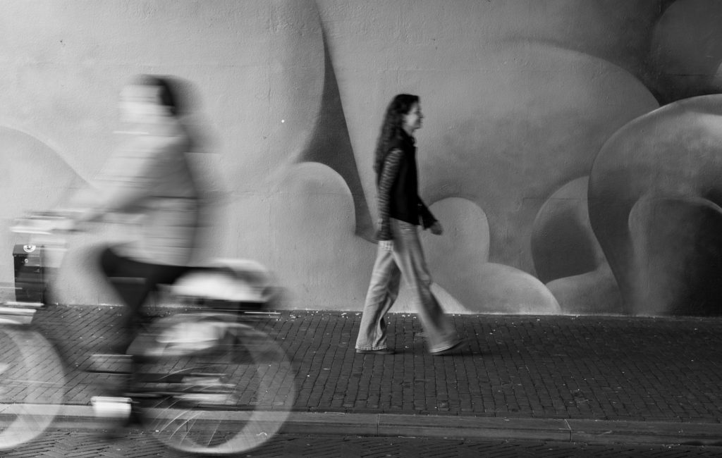 Vrouw loopt onder een brug terwijl een andere vrouw in slowmotion op de fiets voorbijrijdt in een rustige stedelijke omgeving