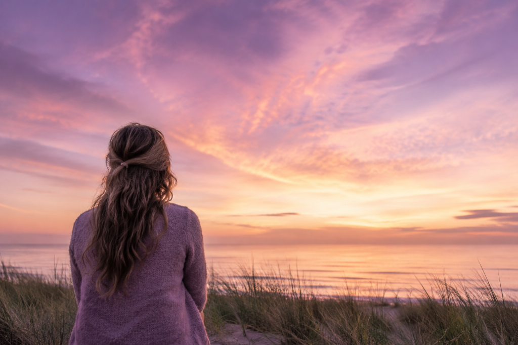 Vrouw kijkt tijdens zonsondergang uit over zee. De zachte paarse, roze en gouden lucht symboliseert rust, liefde en blijvende verbinding na het verlies van een stilgeboren kindje.